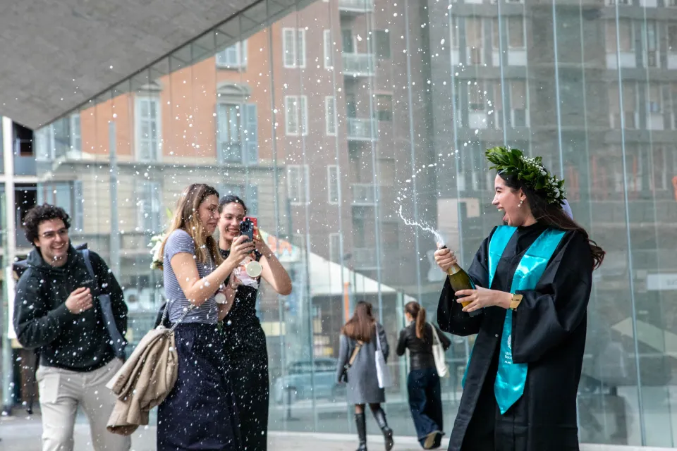 A student celebrates her graduation by uncorking a champagne bottle in front of the Röntgen building.