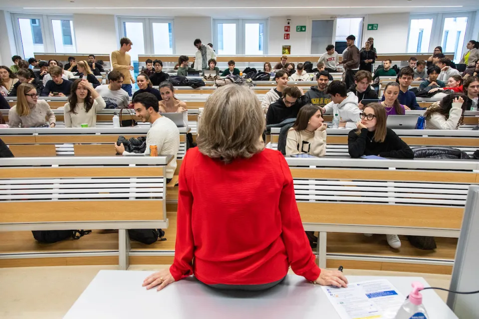 A professor, seen from behind, sits on the edge of the desk watching her students interact before class begins, classroom.