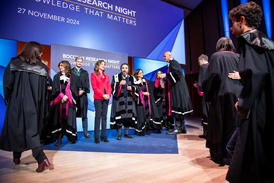 Professors and researchers wearing academic clothes join the Rector on the stage in the Aula Magna in the Röntgen building.